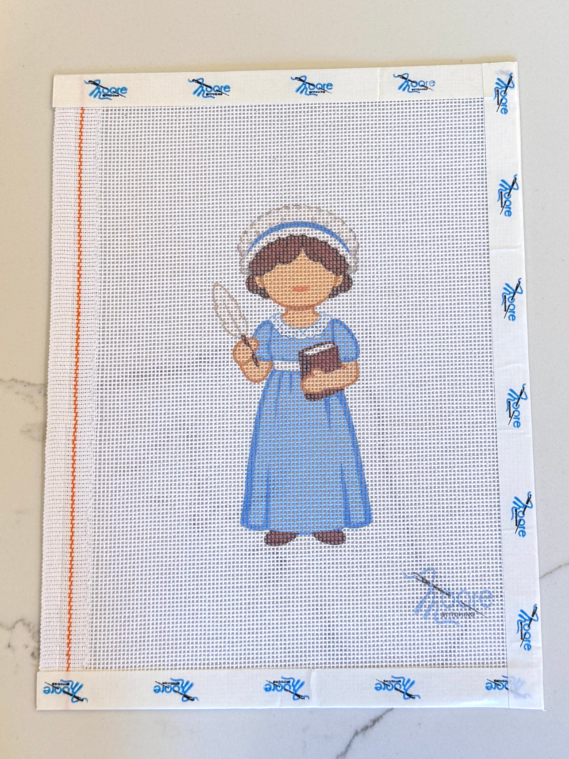 Cross-stitched pattern of a Jane Austen in a blue dress holding a book on fabric with blue border design.