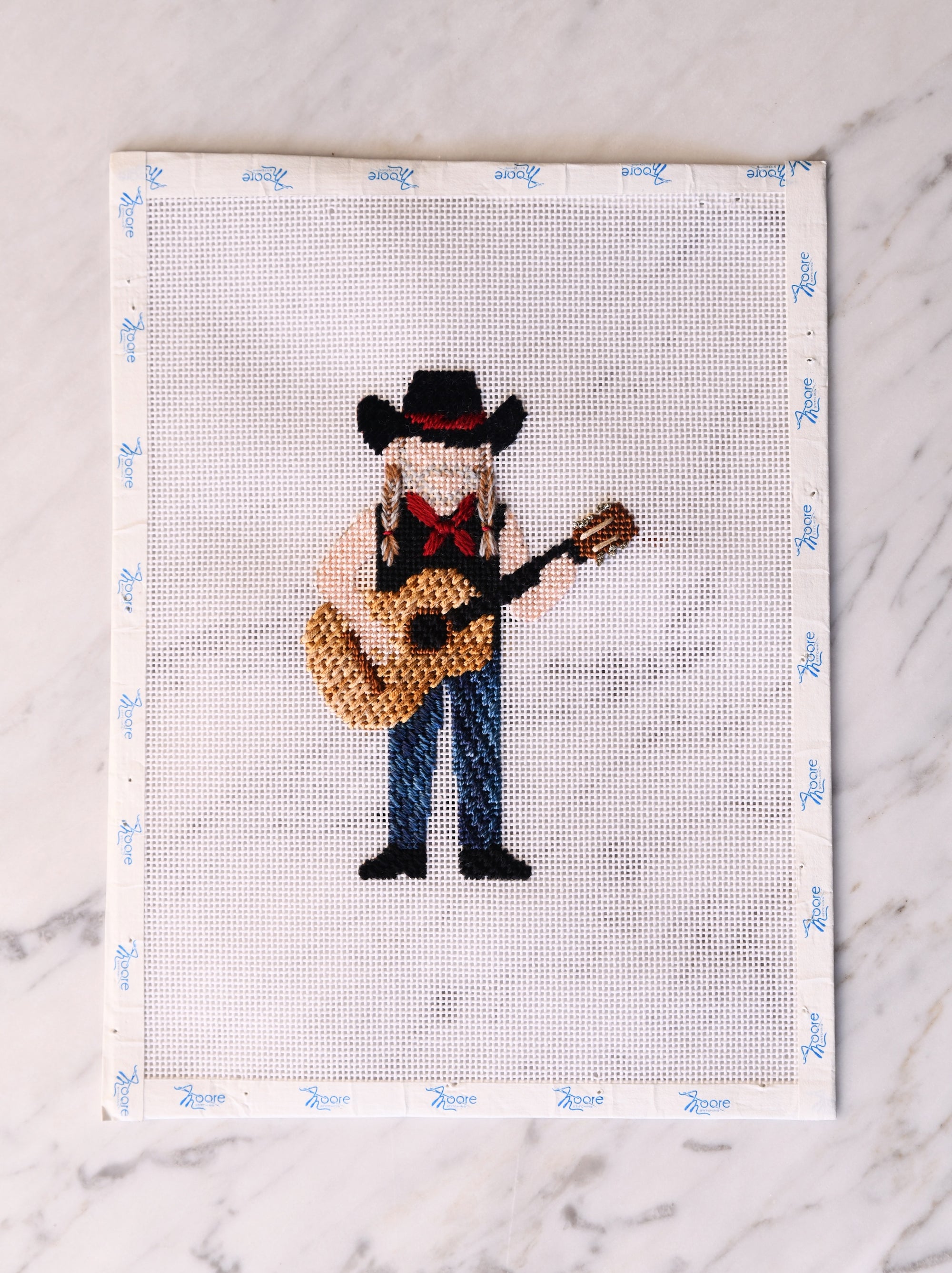 Cross-stitched cowboy playing a guitar on a white fabric background