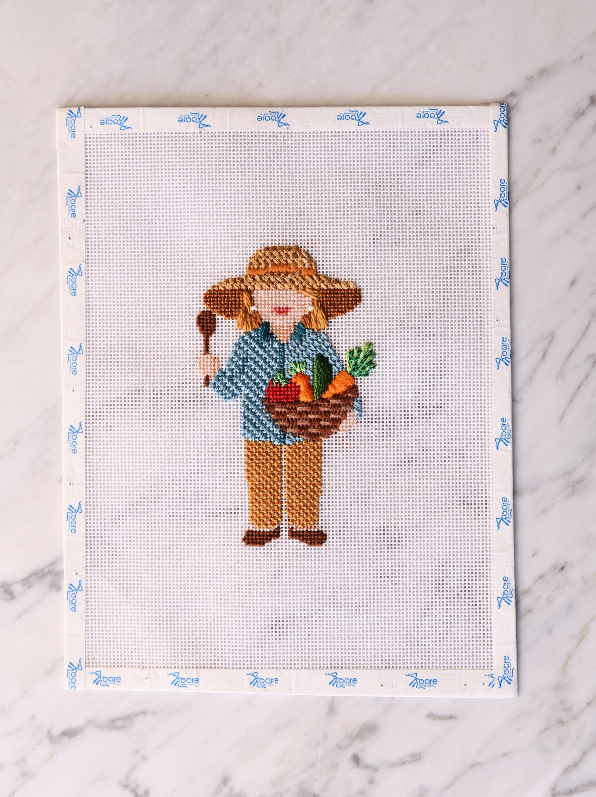 Cross-stitched design of a gardener holding a basket of fruits and vegetables on a white fabric background.