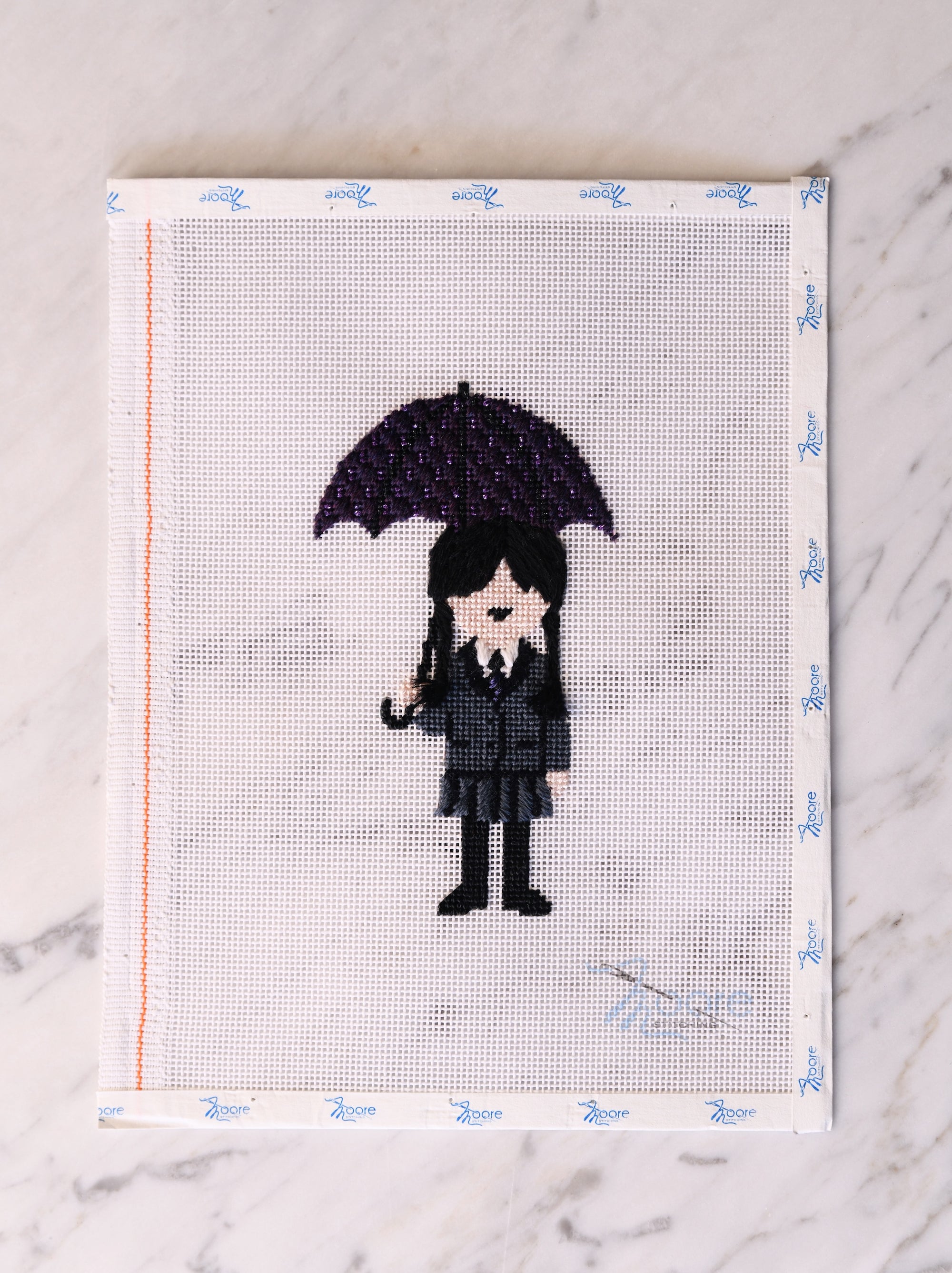 Cross-stitched design of a person holding an umbrella on fabric with a white border.