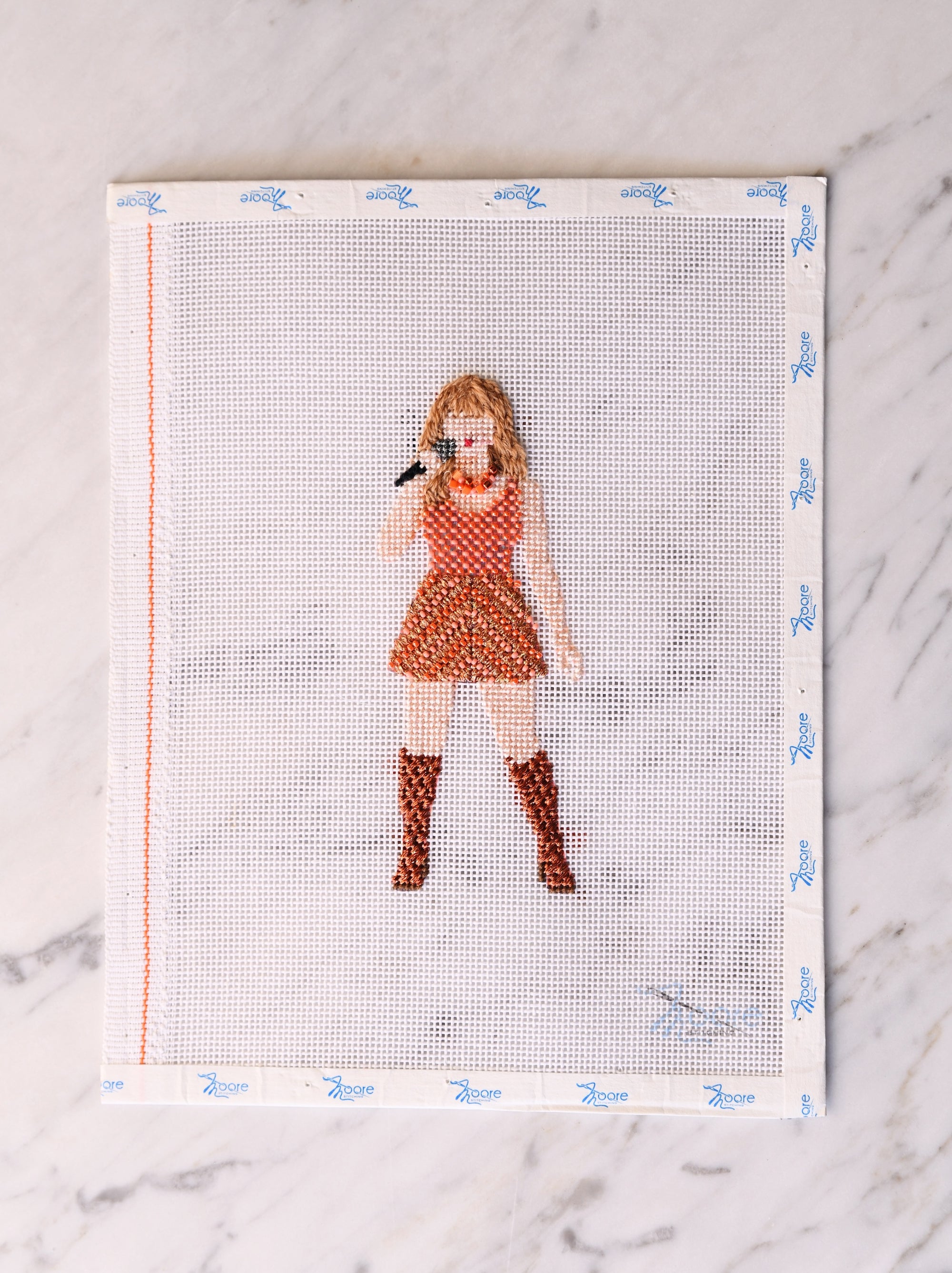 Cross-stitch pattern of a girl in a dress and boots singing in a microphone, on a fabric sheet with a grid.