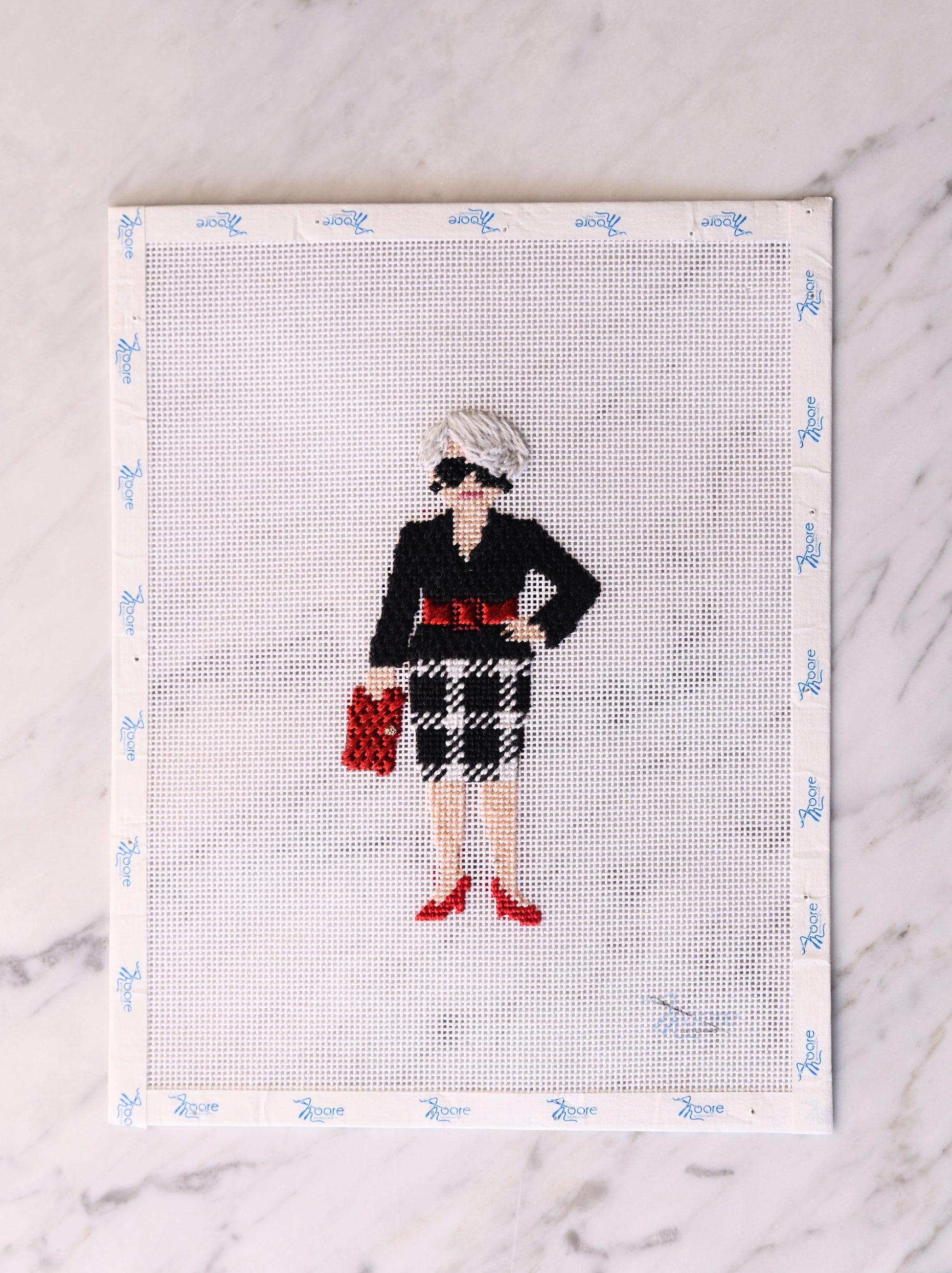 Cross-stitched design of a woman in a black jacket and plaid skirt on fabric with a white border.