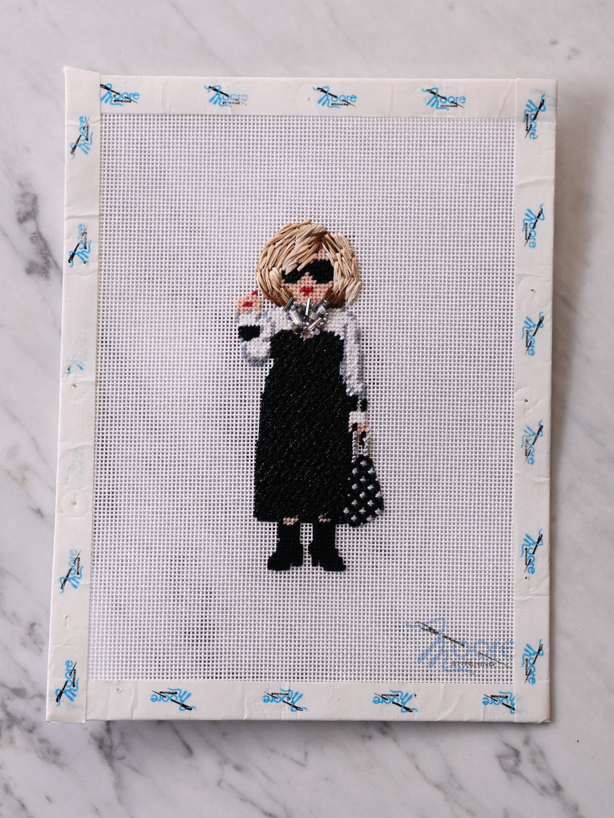 Cross-stitched pattern of a person with a black dress and purse on a fabric sheet.