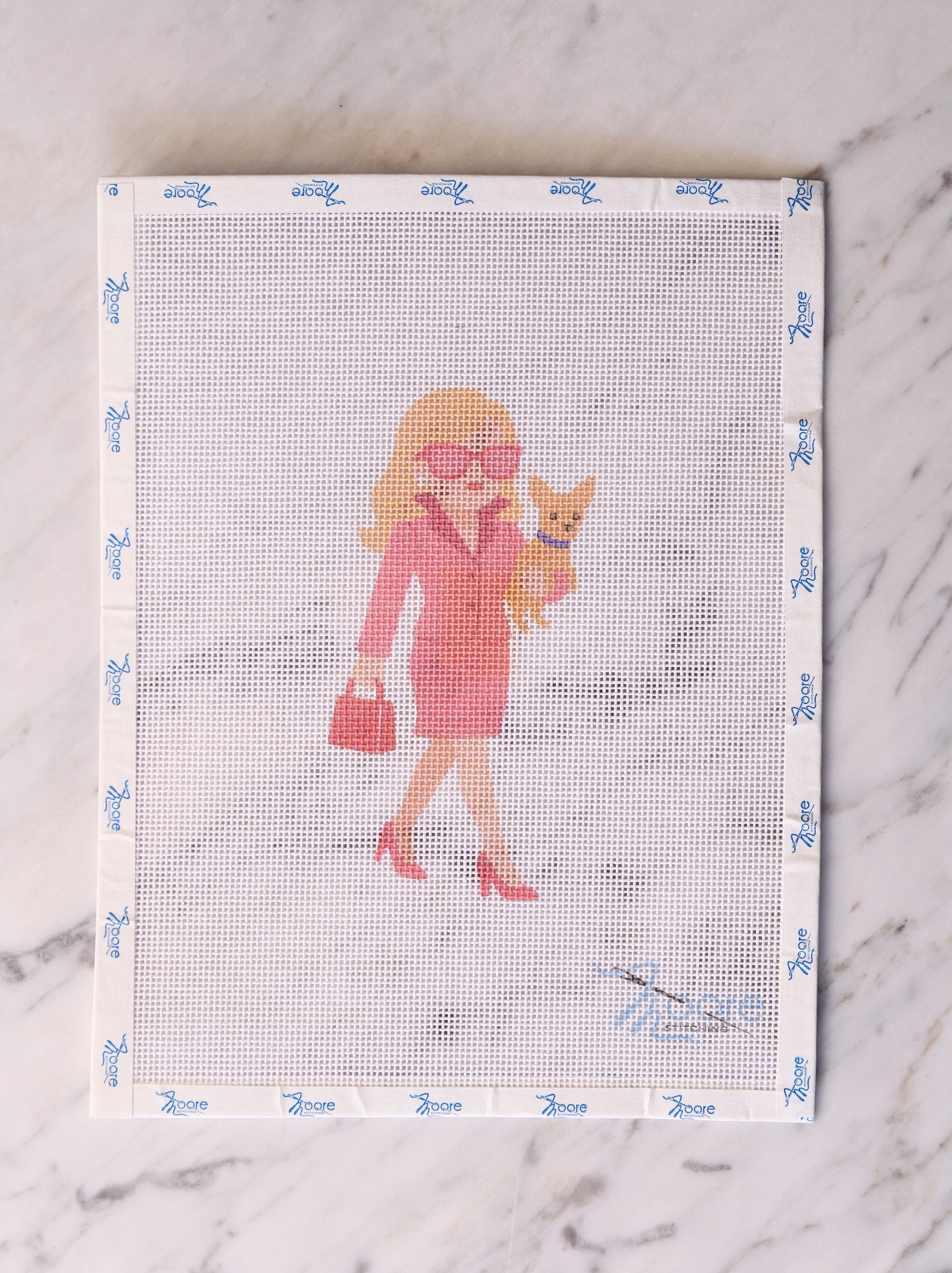 Cross-stitch pattern with a woman in a pink coat and sunglasses holding a dog, on a white fabric background.