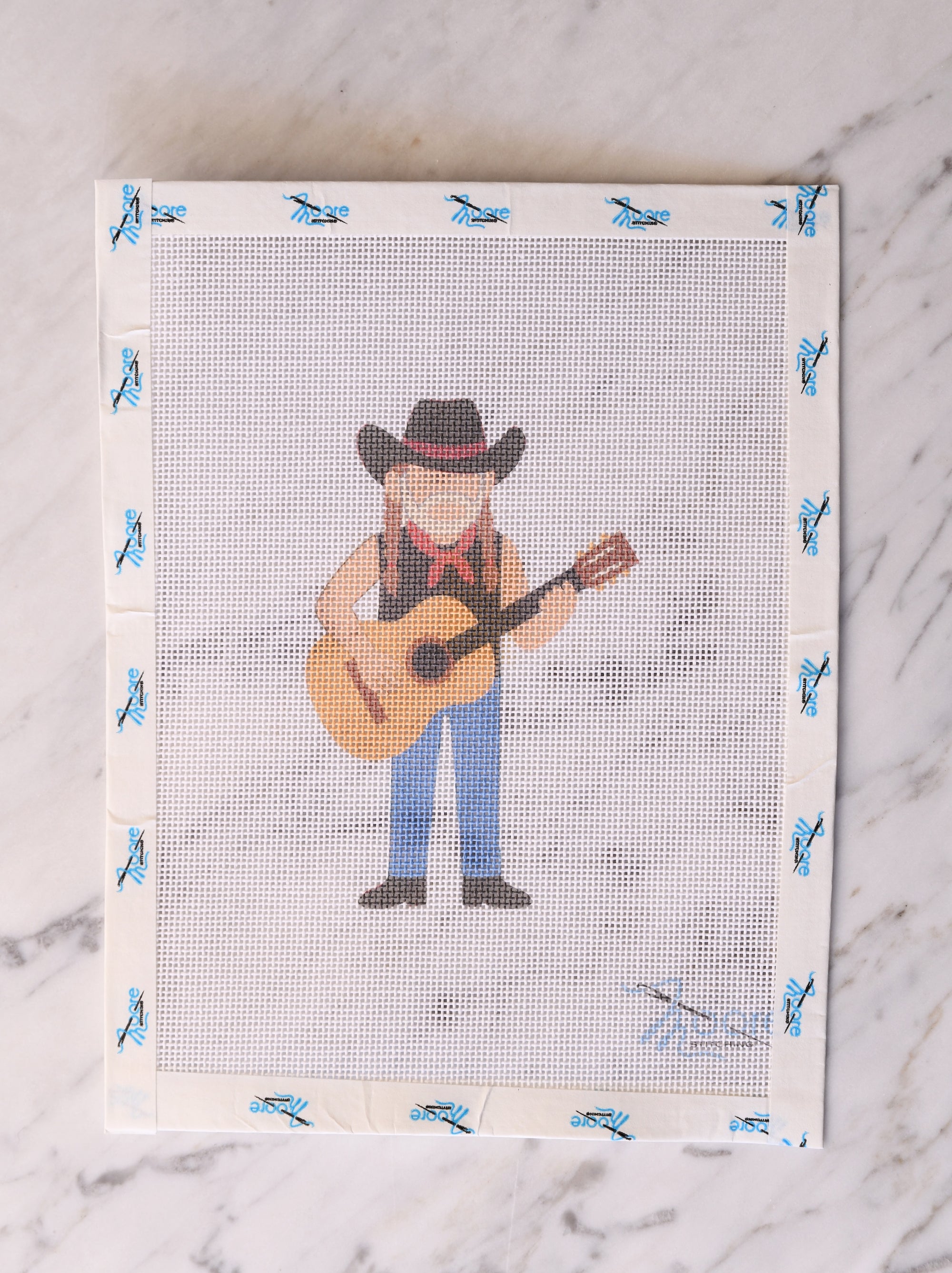 Cross-stitch pattern with a cowboy holding a guitar on a fabric swatch.