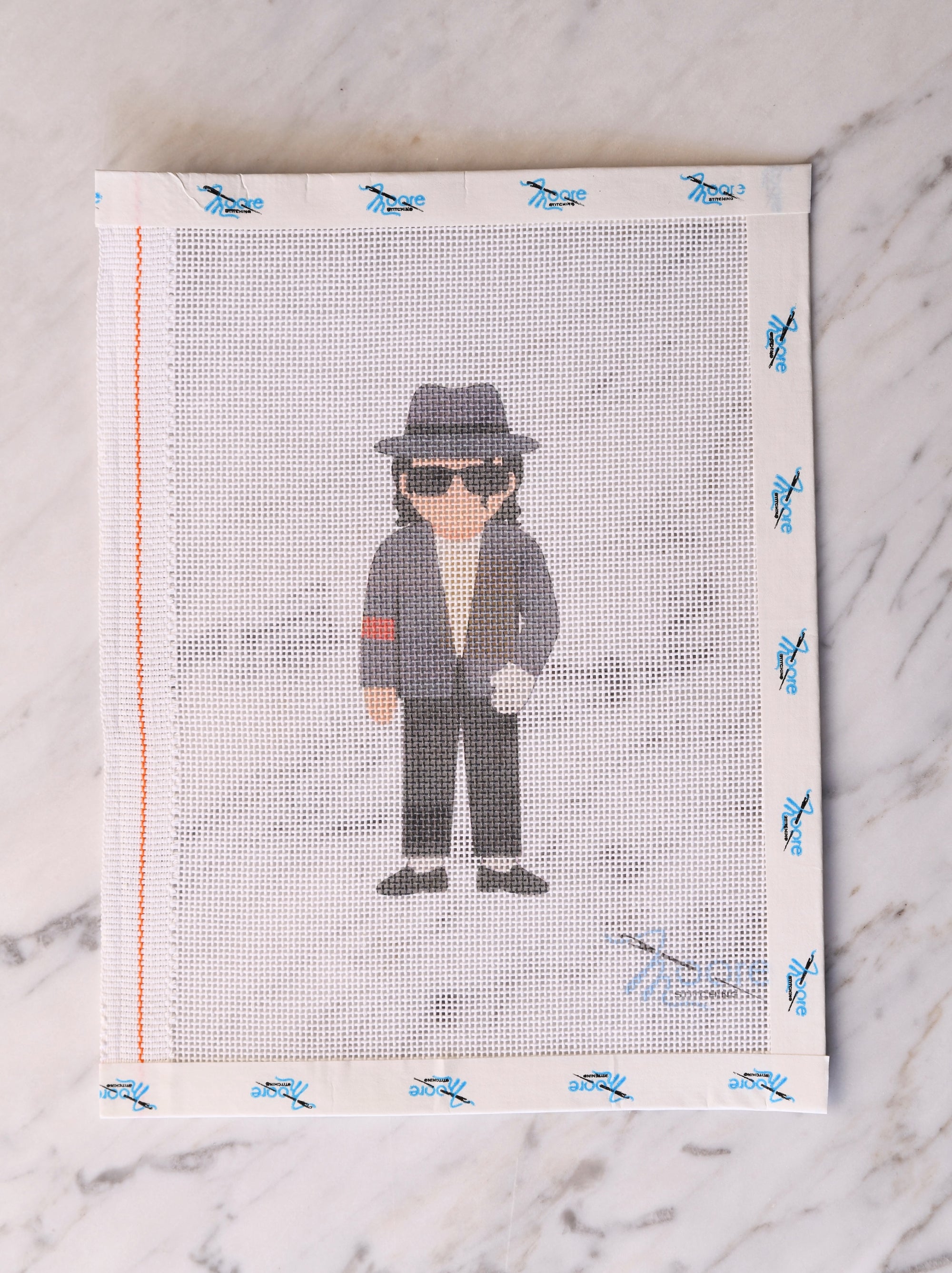 Cross-stitched portrait of a person wearing a hat and sunglasses on mesh fabric.