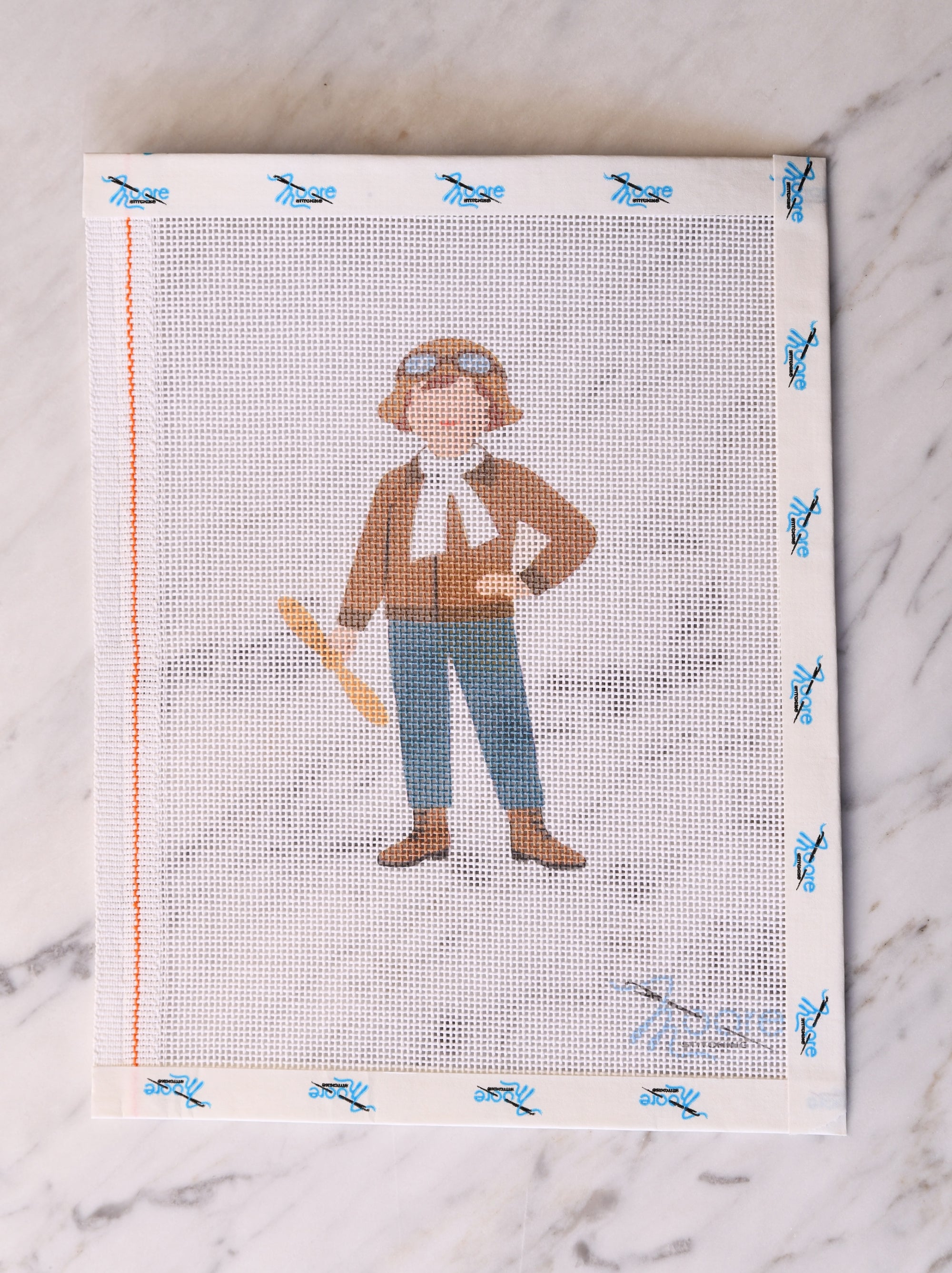 Cross-stitched pattern of a person with a hat and goggles on a fabric swatch.