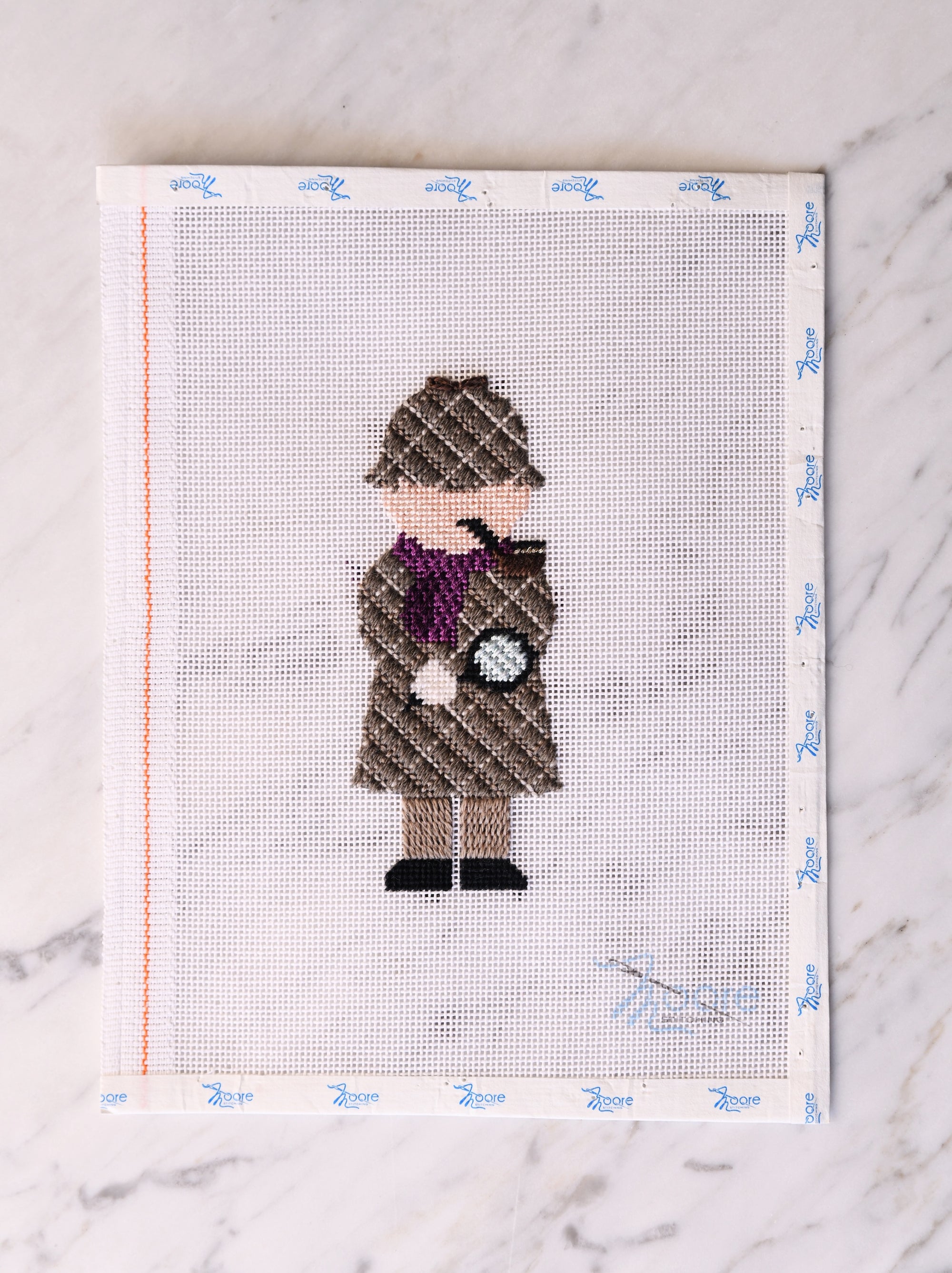 Cross-stitched figure of a detective with a hat and coat on fabric with a grid pattern.