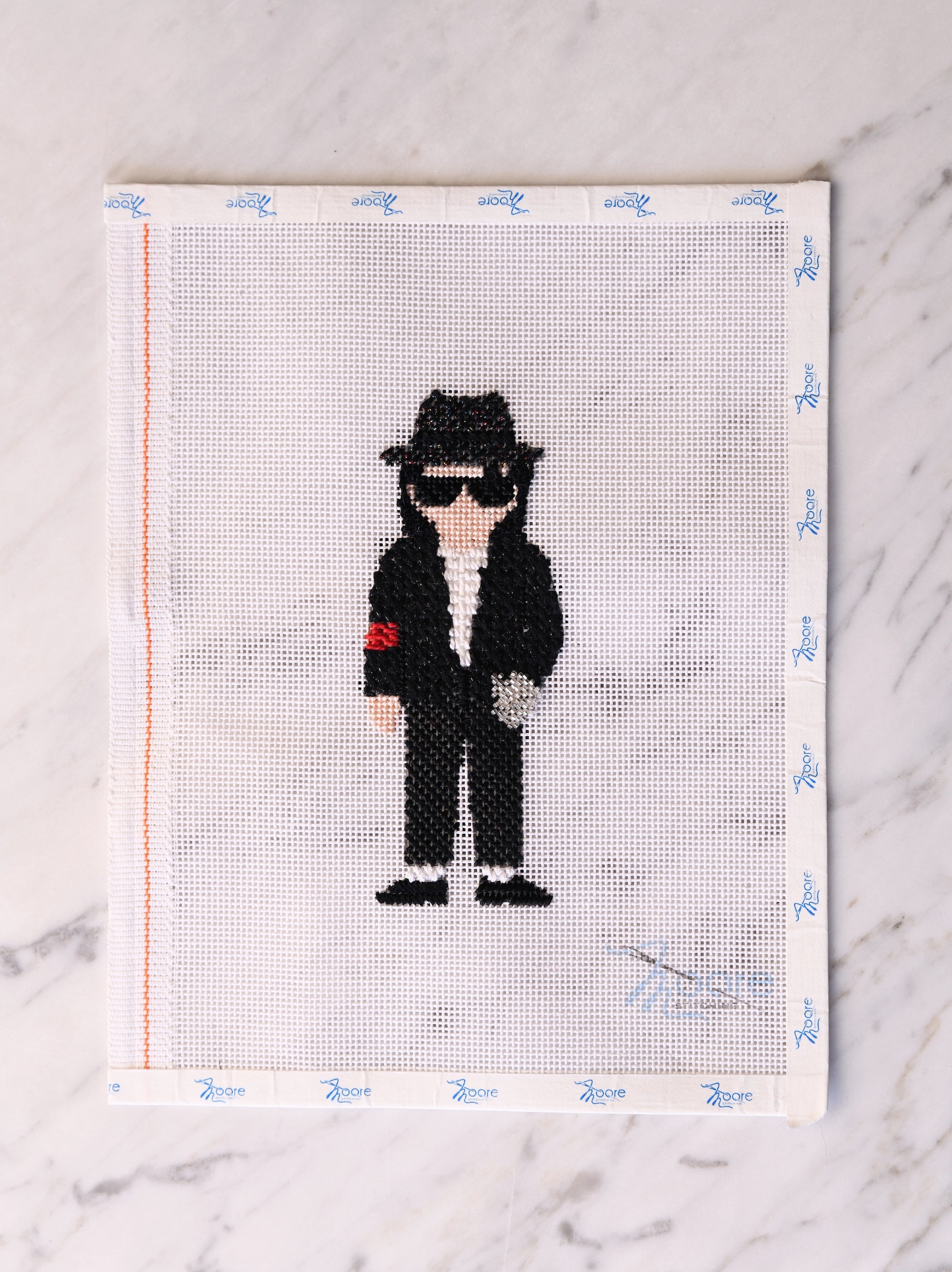 Cross-stitched portrait of a character in a black suit with a silver glove on a white cross-stitching fabric.