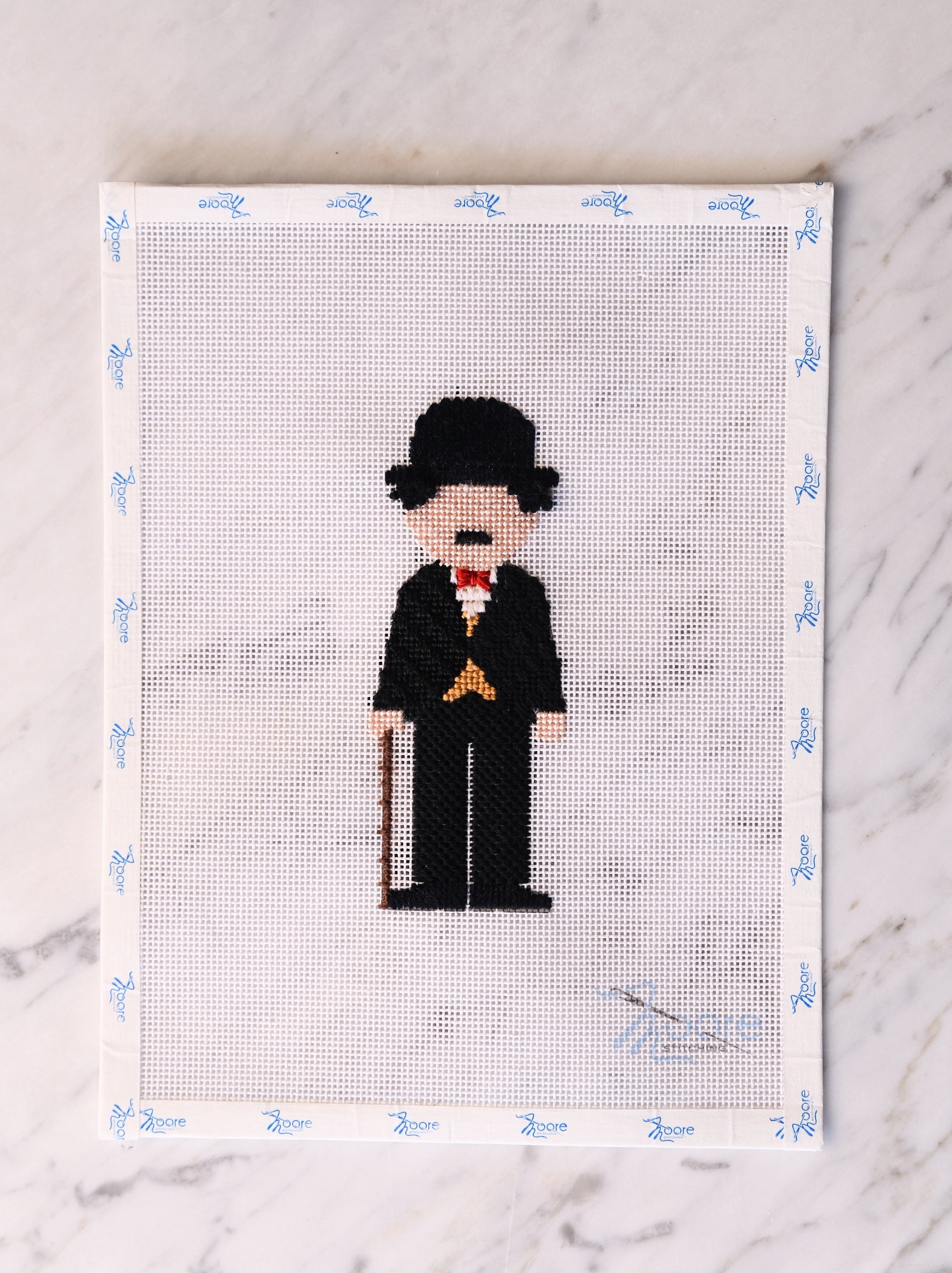 Cross-stitched pattern of a character with a top hat and cane on a white fabric background