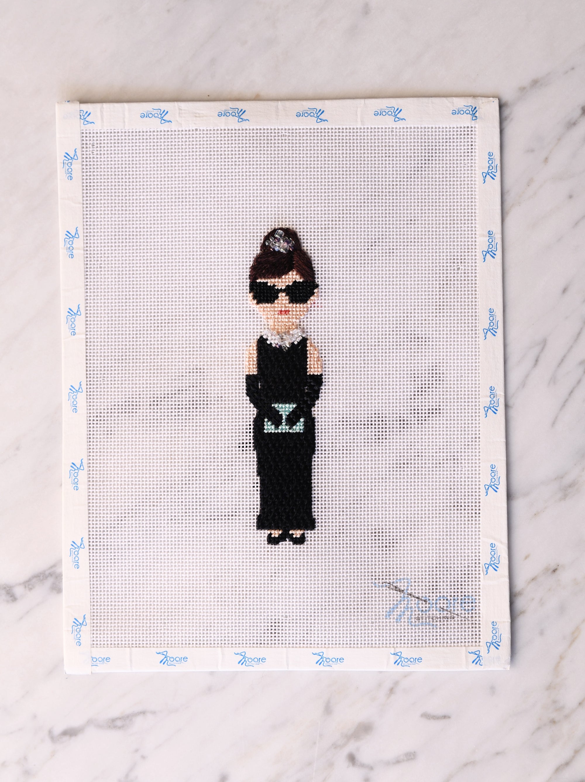 Cross-stitched portrait of a character in a black dress and sunglasses on a white background