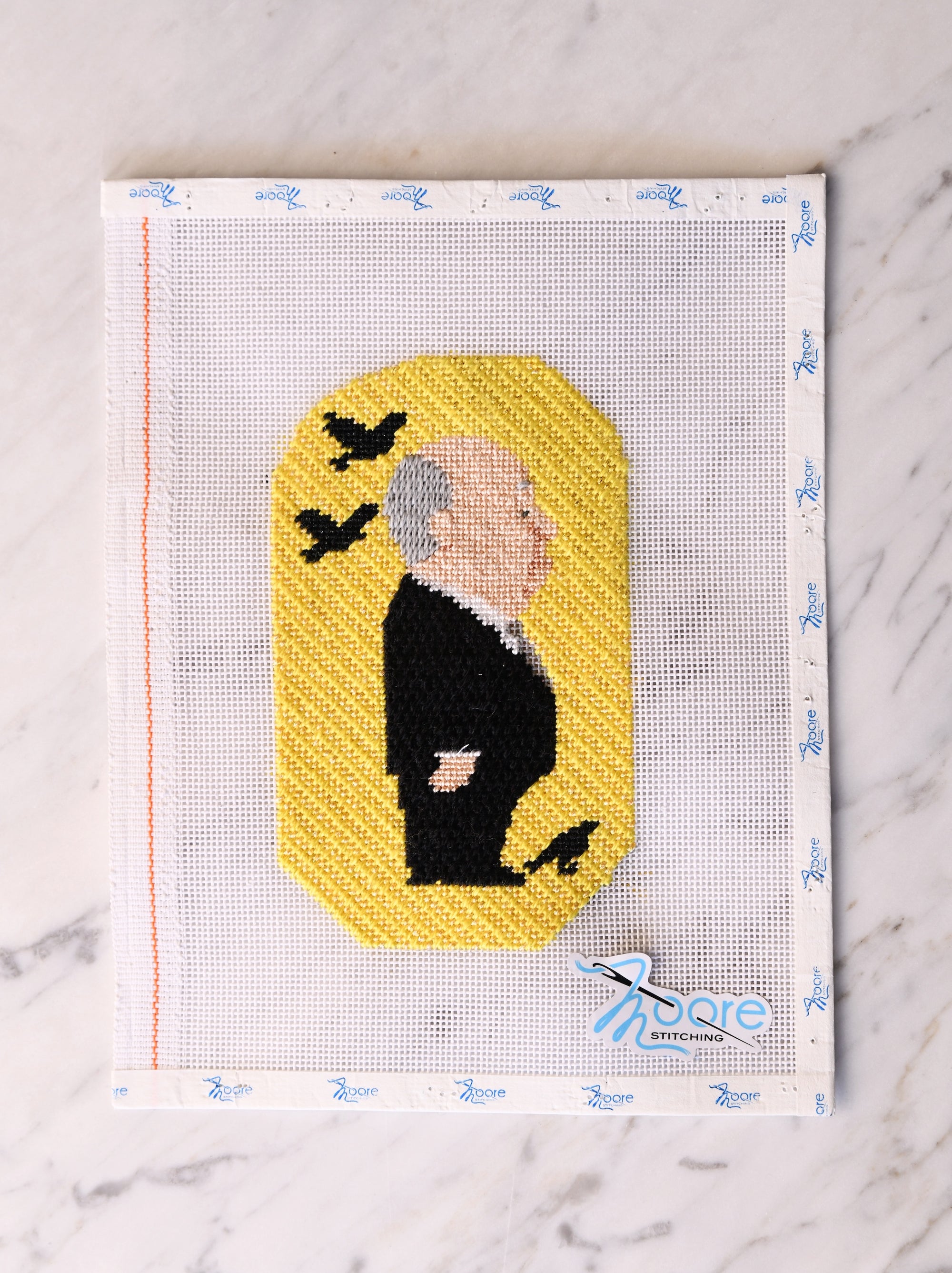 Cross-stitched portrait of a person in a dark suit with birds on a yellow background, on a white fabric.