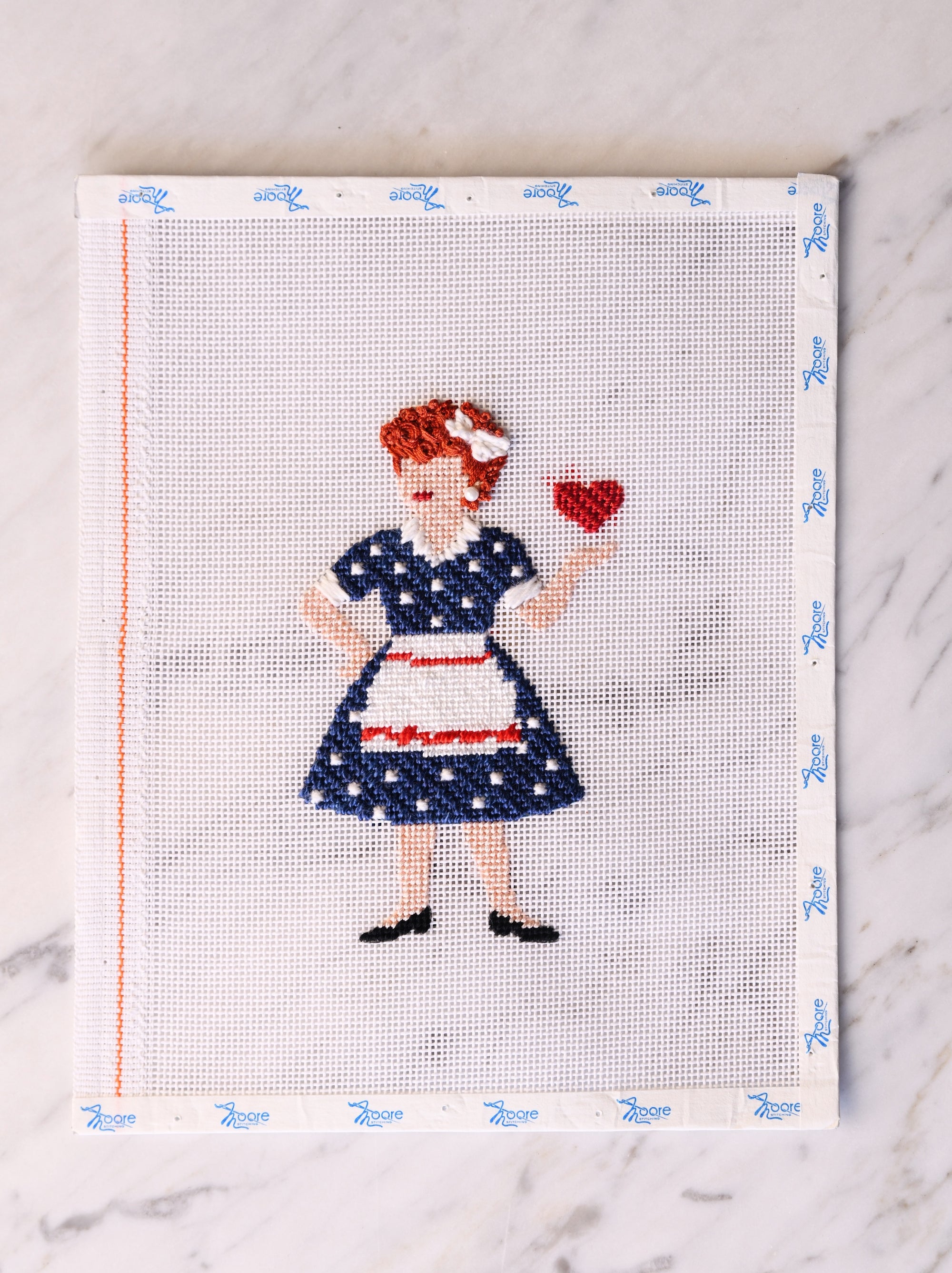 Cross-stitched design of a redhead woman in a polka dot dress holding a heart on a white background.