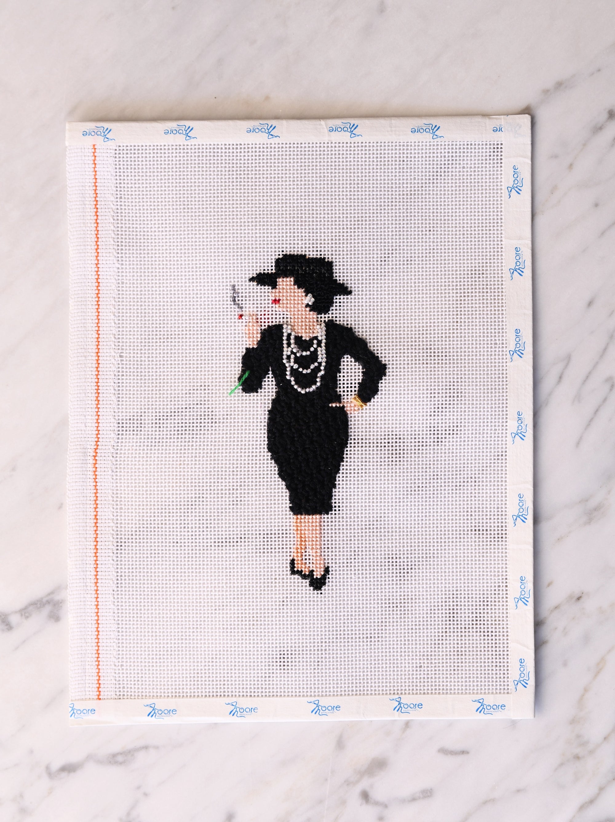 Cross-stitched design of a woman in a black dress and hat on fabric with grid lines.