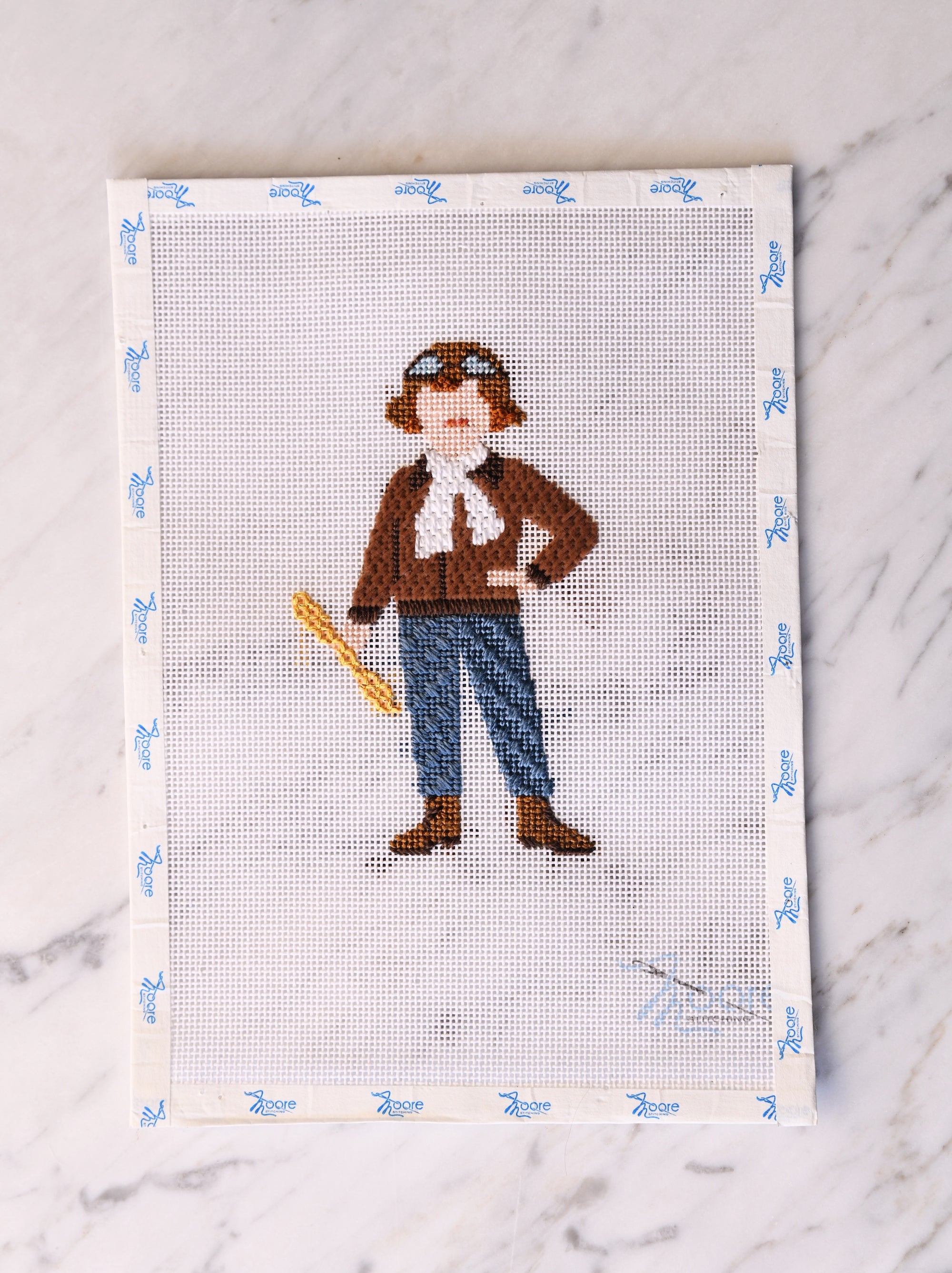 Cross-stitched pattern of a person with a hat, scarf, and goggles on a fabric swatch.