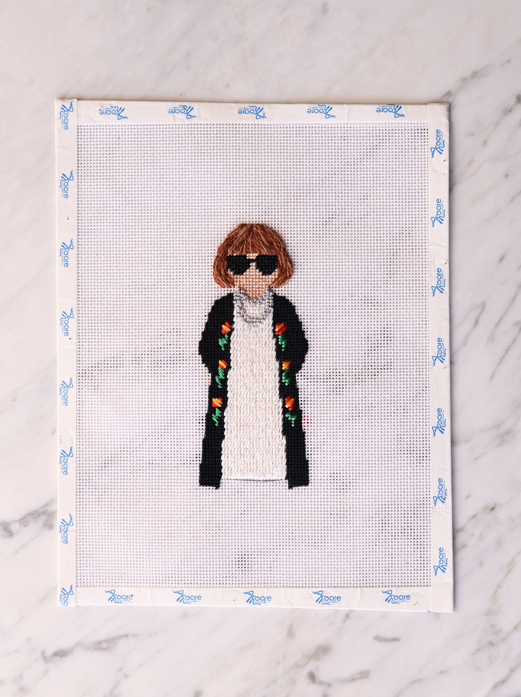 Cross-stitched portrait of a person wearing sunglasses and a coat on a white background