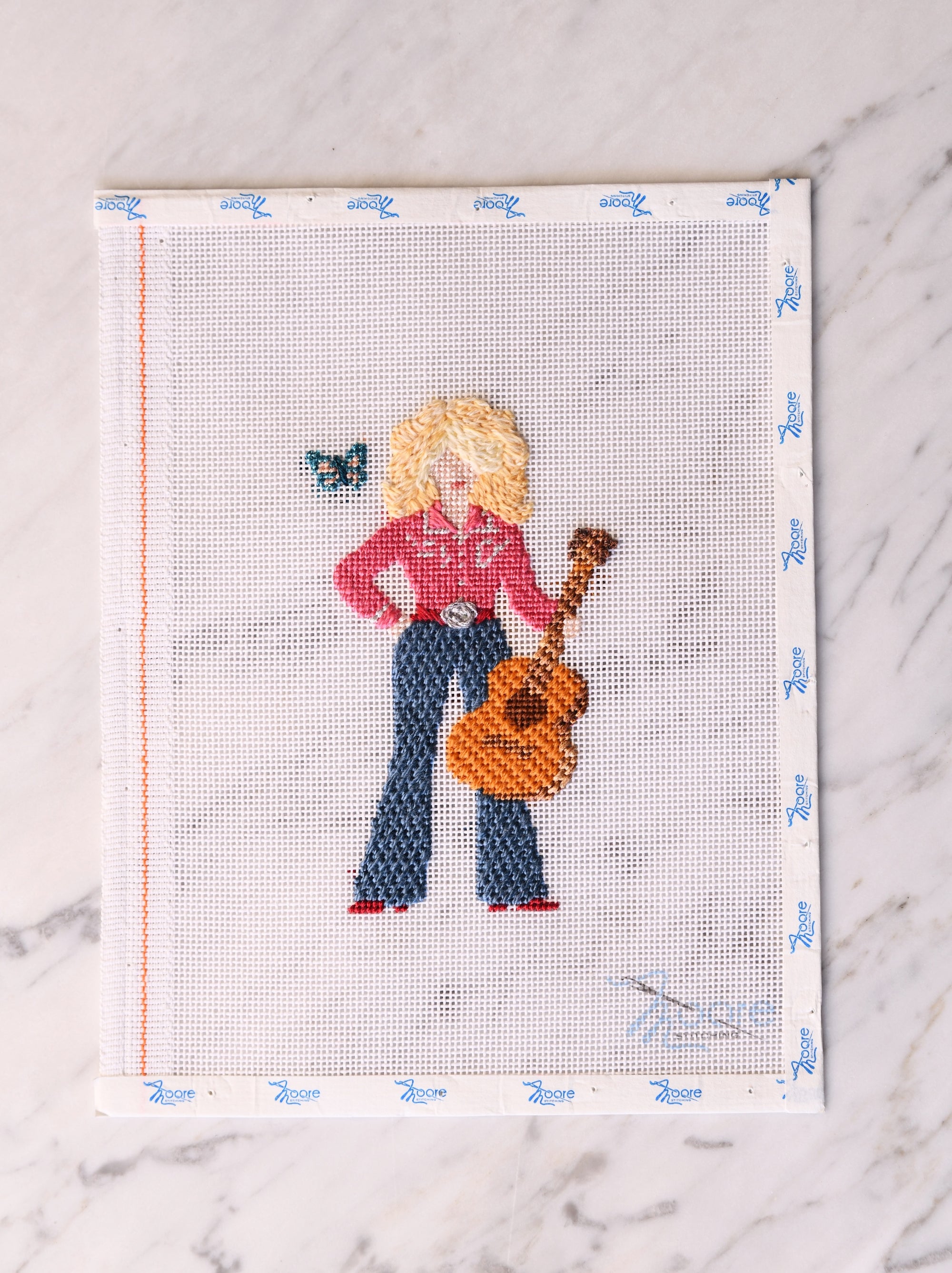 Cross-stitched design of a country girl with a guitar and butterfly on fabric with a white border.